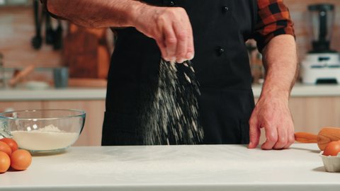 Elderly man preparing food spreading flour in home kitchen. Close up of retired senior chef with uniform sprinkling, sieving sifting raw ingredients by hand baking homemade pizza, bread.