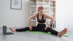 Fitness for beginners. Online training. Home sport. Troubled woman in activewear watching video tutorial on laptop doing hard stretching exercise at light modern room interior.