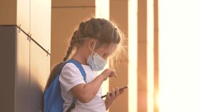 Kid safety mask looks at phone schoolgirl in protective mask.Education during coronavirus pandemic.girl mask uses smartphone.Online education in COVID 19 pandemic.Kid with smartphone online learning.