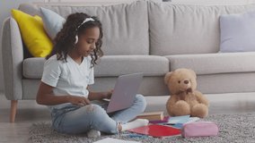 Playtime. Cute little african american girl finish to study online, closing laptop computer and embracing her teddy bear, sitting on floor at home, slow motion
