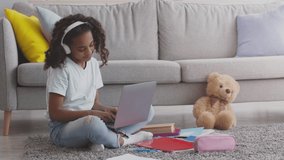 Portrait of happy african american girl typing on laptop, listening to music in headphones, sitting on floor with books and toys at home, slow motion