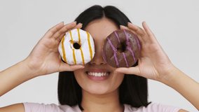 Asian Lady Covering Eyes With Donuts Smiling To Camera Having Fun Standing On White Background In Studio. Unrecognizable Korean Woman Posing With Doughnuts. Sweet Tooth Concept