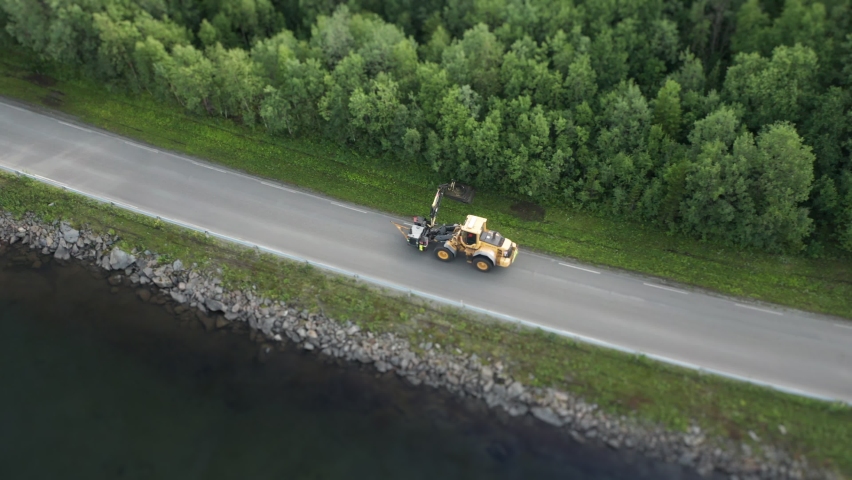 Road maintenance. A bright yellow tractor with a mower is cutting grass, bush and small shrubs on the roadside. The narrow road goes along the bank of the lake. The lush forest is on the other side.