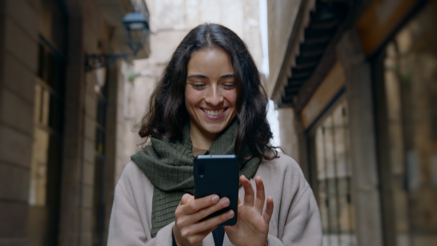 Young smiling dark-haired woman wearing light-colored coat, walking city streets and using mobile device to chat with her friends. Good vibes. Modern technology usability concept. Slow motion shot Royalty-Free Stock Footage #1074328412