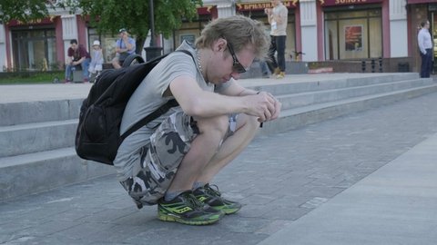 RUSSIA, NIZHNIY NOVGOROD, 18 MAY 2021: young man in grey shirt, shorts and sunglasses sitting at pavement, using smartphone. caucasian male takes photographs or watching series on his phone
