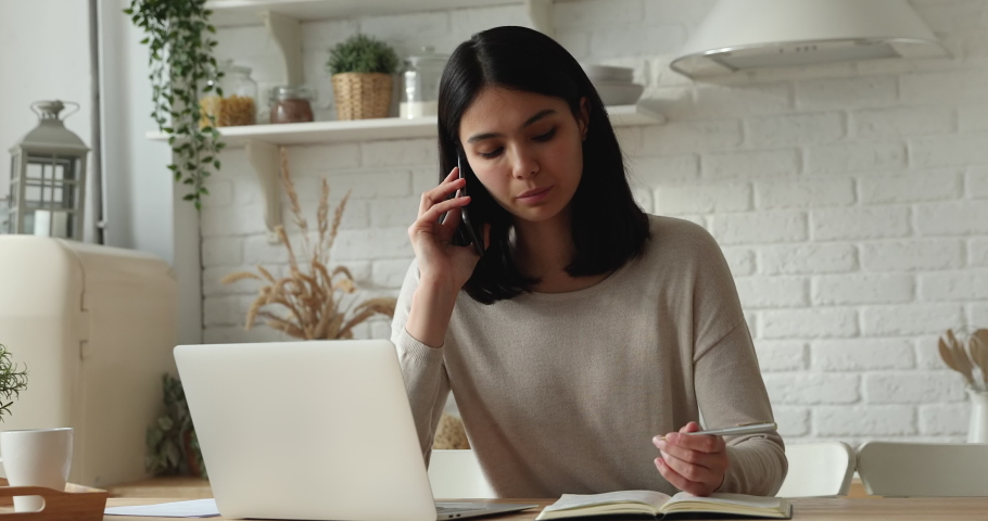 Young Asian ethnicity woman text on laptop talks on phone make work call, communicates with colleague remotely while sitting in kitchen at home. Telesales occupation sell services by telephone concept Royalty-Free Stock Footage #1074328940