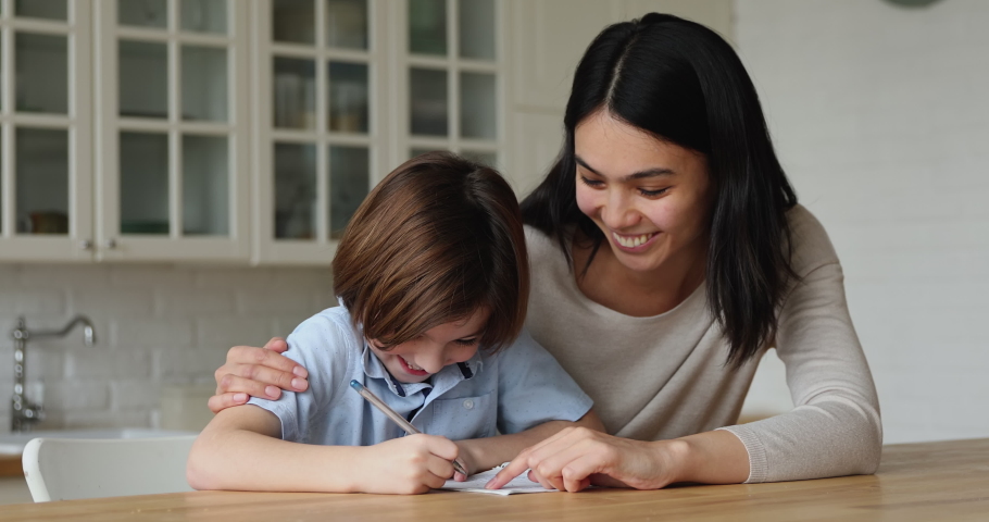 Korean mom helps to son with homework, multi ethnic family sit in kitchen schoolkid takes notes in copybook accomplish task receives from tutor praises, giving high five gesture, homeschooling concept Royalty-Free Stock Footage #1074328961
