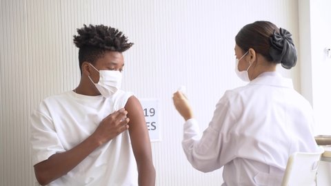 Portrait of African-American man looking at while getting covid vaccine in clinic or hospital, with hand nurse injecting vaccine to get immunity for protect virus. teenager wearing protective mask.