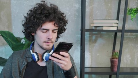 Young Caucasian man sit at desk at home office and talk on loudspeaker on smartphone. Millennial male use cellphone to record voice message.