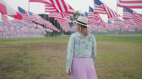 Woman walking between waving American flags in park. 4 of July, Independence day celebration 4K footage. Slow motion back view camera following female. Patriotic holiday, democracy and veteran respect