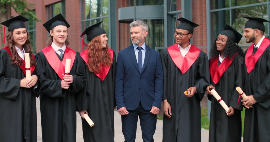 This is final. Full length view of the group of students at the graduation gowns and caps standing near the university with their teacher and showing their diplomas to the camera