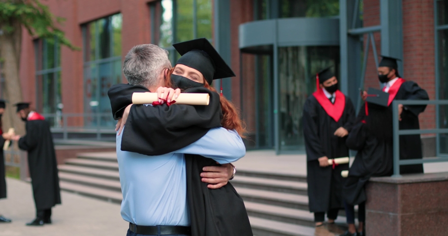 Happy father and his student daughter wearing protective mask on graduation day embracing with each other. Girl in academic gown and cap hugging her father in front of the camera Royalty-Free Stock Footage #1074337220