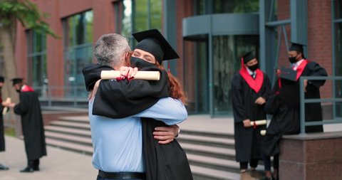 Happy father and his student daughter wearing protective mask on graduation day embracing with each other. Girl in academic gown and cap hugging her father in front of the camera