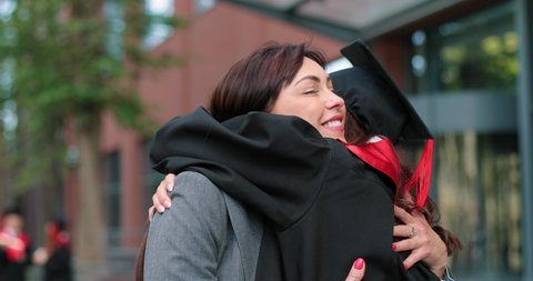 Happy female graduate are embracing with her mother with diploma at her hands and rejoicing with each other. Woman is hugging her daughter with other students moving and talking in background