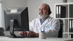 Grey male doctor remotely video communications with customer and records medical history in computer
