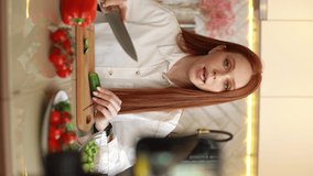 Tracking shot of female vlogger shooting video food blog about cooking on camera of mobile phone at table in kitchen. Young woman blogger holding knife cutting fresh cucumber for vegetable salad.