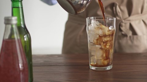 Man in apron slowly pours coffee into a tall glass filled with ice cubes. Coffee is poured into a glass with ice. Chilled drink. Preparing coffee summer ice coffee in the morning.