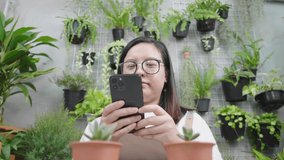 Front view of happy asian woman wearing glasses taking a picture of plants and sending to her customer by smartphone, sitting in the garden at home. Chatting with customer. Selling online concept