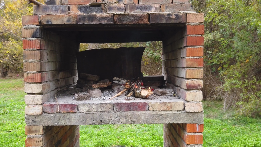 Fire in outdoor bbq fireplace, made from bricks among a green meadow and trees. White smoke on an autumn day in the forest.