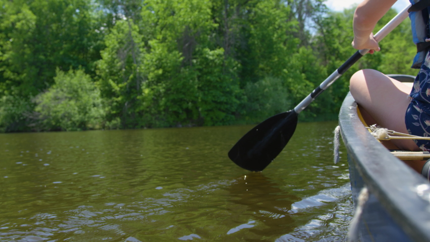Woman rowing a canoe down a beautiful river surrounded by lush trees. Low angle shot, slow motion.