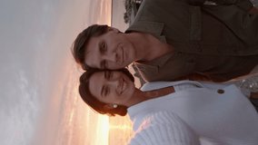 Vertical view shot of young Caucasian man and his beloved bride with camera in her hand are standing together on sea coast at sunset and making selfies