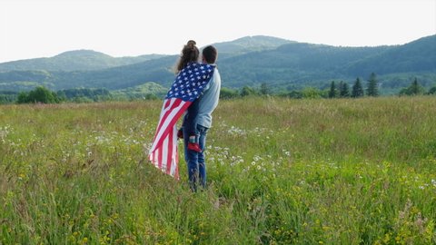 Back view of young American father holds little daughter wrapped in USA flag standing on beautiful meadow on background mountains, enjoying amazing summer nature view on sunny day. July fourth concept