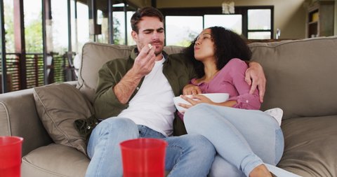 Smiling mixed race couple eating popcorn sitting together on the couch at vacation home. couple honeymoon and vacation concept