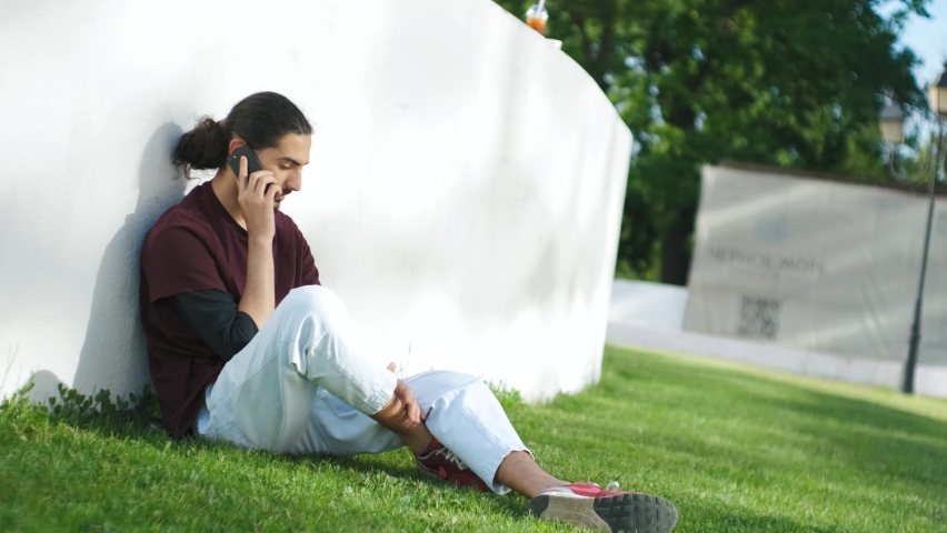 portrait of young attractive arab man sitting on the grass and talking on the phone. Man in park