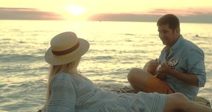 Fun attractive couple of traveler sitting on sea beach with ukulele under sunset sky in evening time. Man playing, woman listening and sing music, relaxing enjoying holidays, travel day. 4K video
