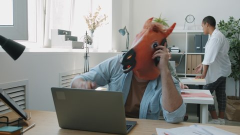 Angry businessman wearing horse mask is talking on mobile phone and gesturing sitting at desk in office while coworkers are having fun in background