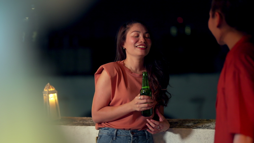 Asian woman friends drinking beer from a bottle with talking together with happiness. Smiling millennial people female friends enjoy dinner party at outdoor rooftop for meeting reunion at night