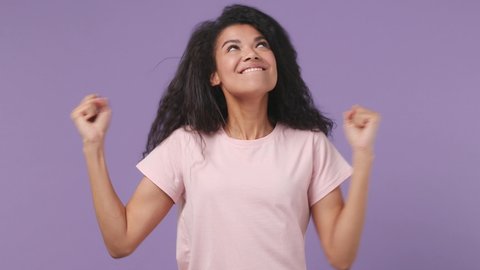Excited jubilant overjoyed young african woman 20s years old wears pink T-shirt doing winner gesture celebrate clenching fists say yes isolated on pastel violet purple color background studio portrait