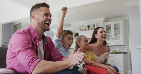 Excited caucasian parents, son and daughter on couch watching tv and cheering, son holding football. happy family, at home together.