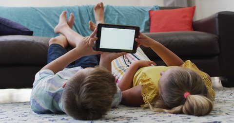 Happy caucasian brother and sister lying on floor in living room using tablet, copy space on screen. happy family, free time at home.
