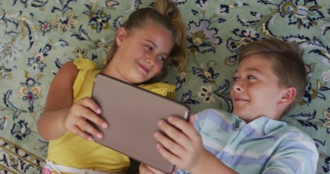 Happy caucasian brother and sister at home, lying on floor using tablet together and smiling. happy family, free time at home.