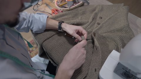 Top view of male tailor sewing button on knitted jacket. Professional man working in atelier studio indoors. Concentrated dressmaker stitching clothes