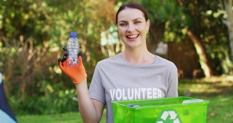Smiling caucasian woman wearing volunteer t shirt holding recycling crate, collecting plastic waste. eco conservation volunteers doing countryside clean-up.