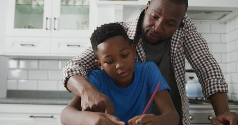 Happy african american father leaning over son sitting at kitchen table helping with his school work. family spending time together at home.