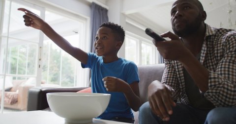 Happy african american father and excited son watching tv and eating popcorn in living room. family spending time together at home.