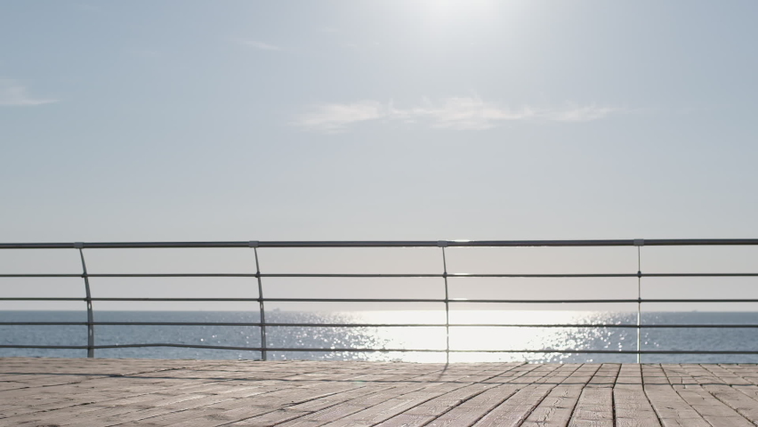 Lady rides electric scooter along brown wooden boardwalk and guy runs following against endless blue sea super slow motion