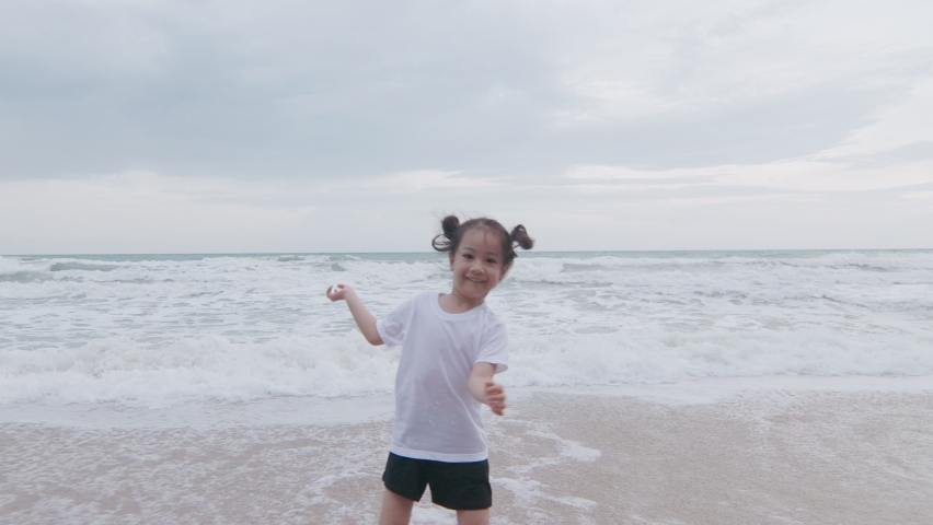 4K of adorable 5 years old asian girl with black hair is running with happy smile and enjoyment near ocean coast seeing strong wave and horizon for sunset which is relaxing holiday for kid and family.