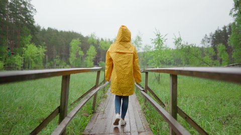Back view follow shot of young woman in yellow raincoat with hood walking down wooden path in countryside. Female traveling in woodland in rain