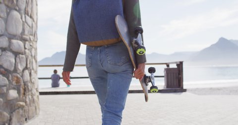 Rear view midsection of mixed race woman holding skateboard walking on promenade by the sea. healthy living, off the grid and close to nature.