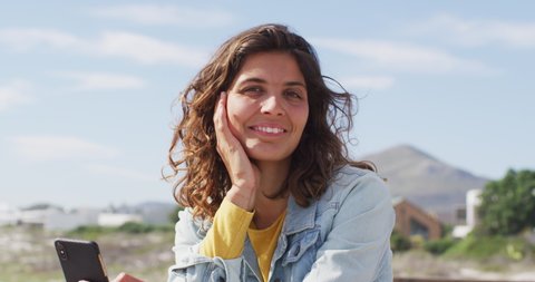 Portrait of happy mixed race woman holding smartphone and smiling on sunny promenade by the sea. leisure time, outdoors and close to nature.