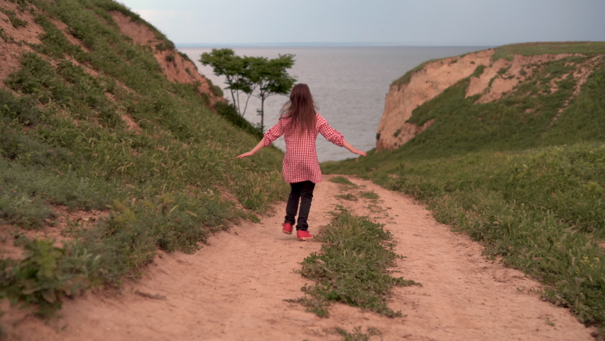 A little girl spreads her arms and runs away on the road towards the sea or ocean. Freedom and relaxation concept. The child is resting.