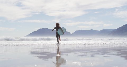 Rear view of mixed race woman running from beach into the sea carrying surfboard. healthy active lifestyle, close to nature.