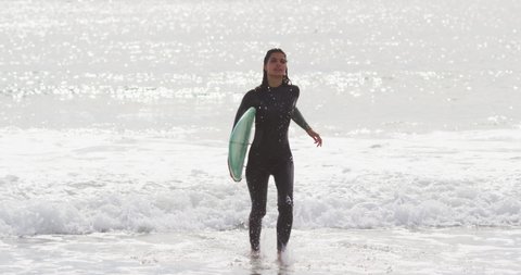 Mixed race woman running from the sea onto beach carrying surfboard. healthy active lifestyle, close to nature.