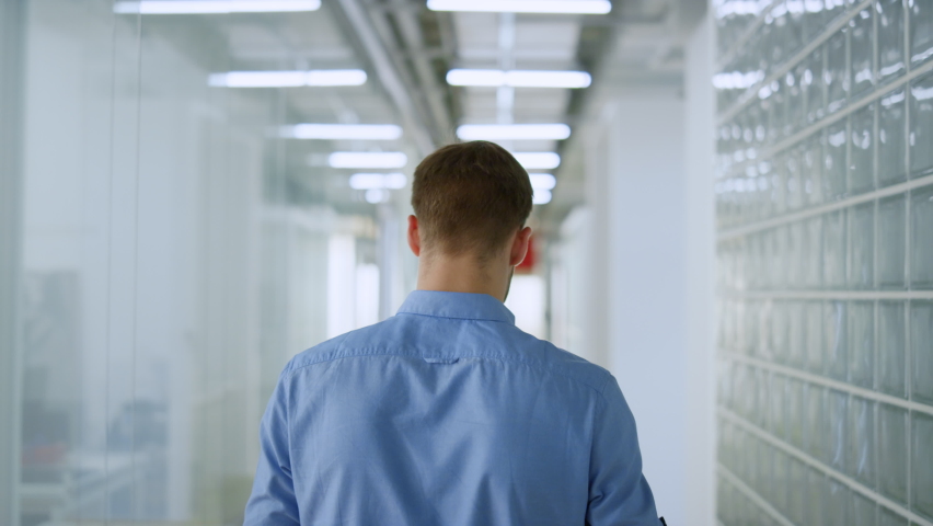 Unknown young man walking through trendy office. Back view businessman looking around in modern interior. Unrecognizable business male person going along glass wall space. Royalty-Free Stock Footage #1074408479