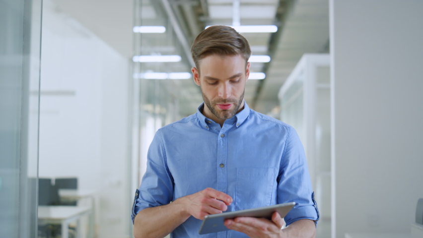 Joyful business man is using tablet computer while walking on modern office. Confident businessman looking digital device screen in modern interior. Young entrepreneur working indoors.  Royalty-Free Stock Footage #1074408482