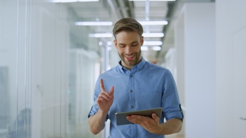 Joyful business man is using tablet computer while walking on modern office. Confident businessman looking digital device screen in modern interior. Young entrepreneur working indoors. 